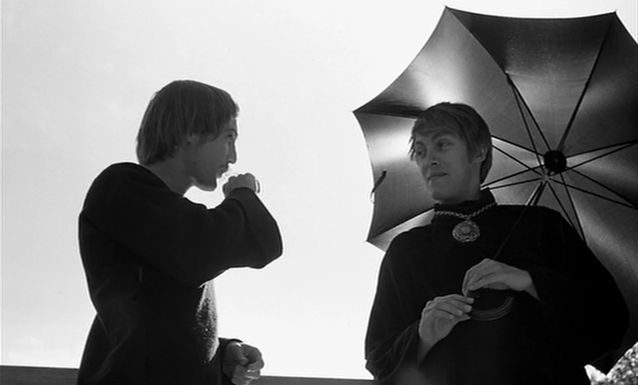 Stereo (1969) movie still: medium — Two individuals stand in an open outdoor setting against a bright, overexposed sky. One figure holds a large…