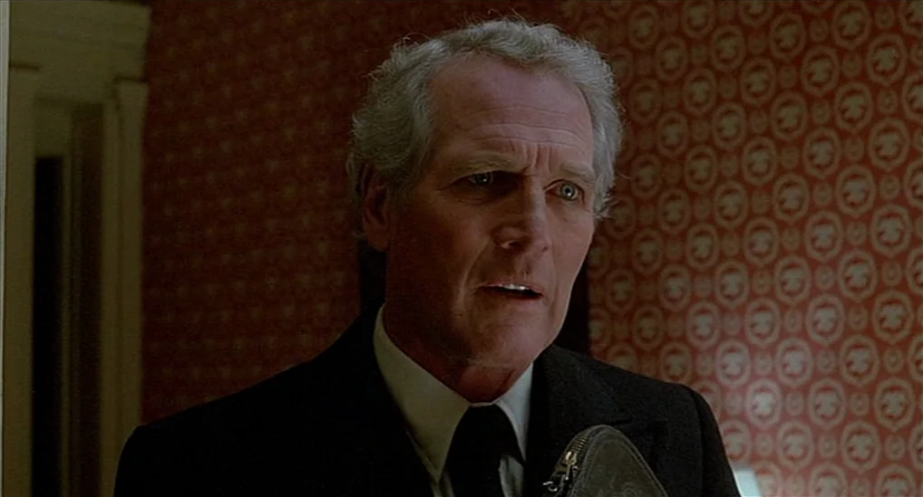 The Verdict (1982) movie still: closeup — A man stands in a hallway decorated with classic red floral patterned wallpaper. He is wearing a formal…