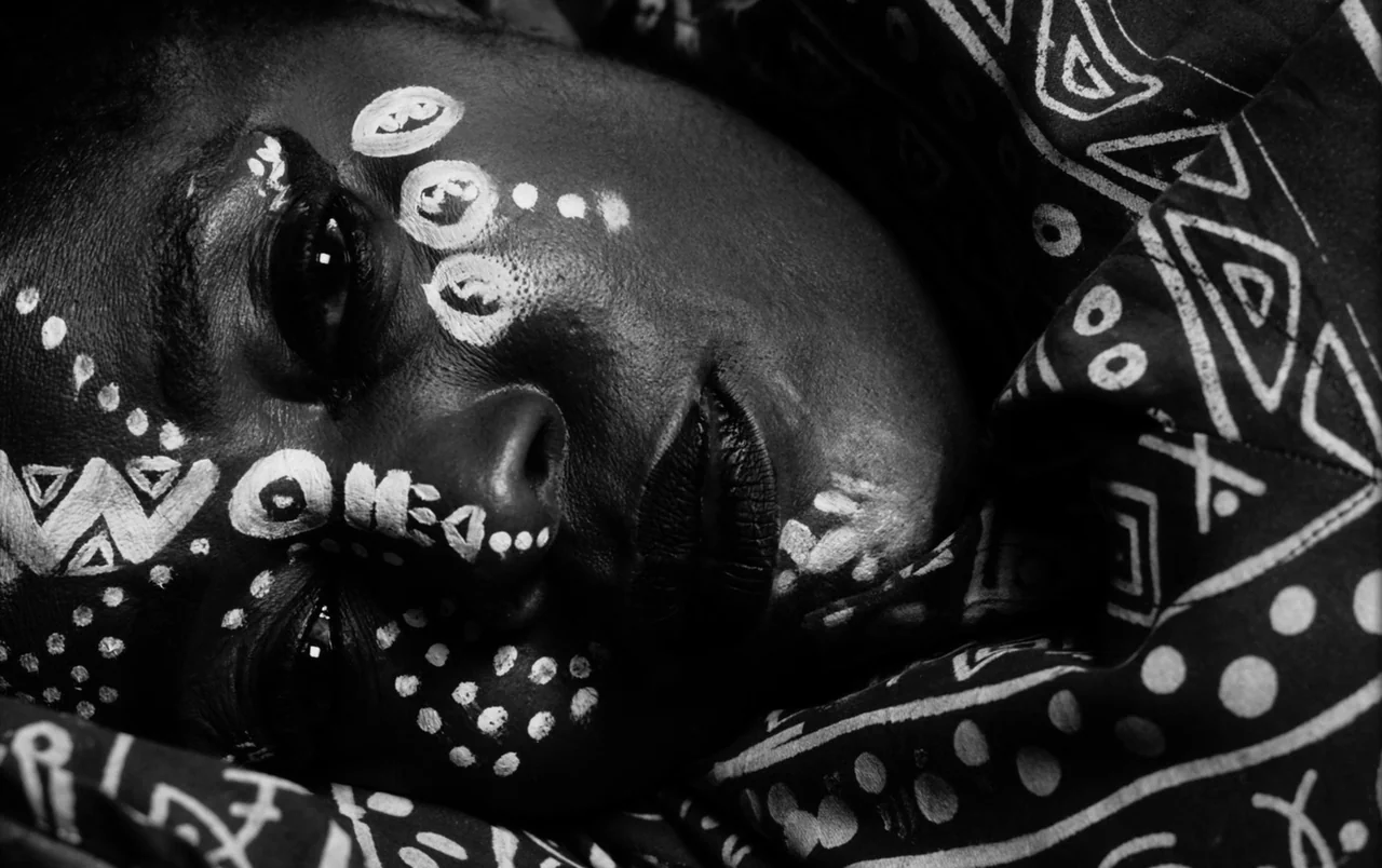 Mami Wata (2023) movie still: closeup — A close portrait shows a person with intricate white painted designs on their face. The subject rests their…