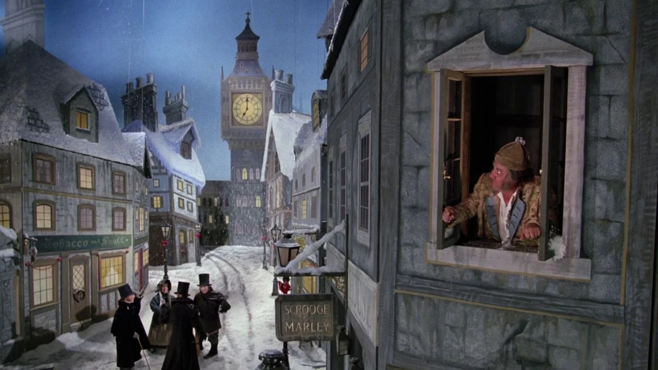 Scrooged (1988) movie still: wide — A theatrical set designed to look like a snowy Victorian town square with painted backdrops. A man leans out of…
