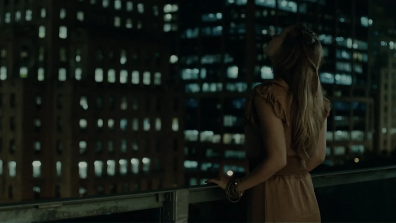 The Words (2012) movie still: medium — A dark balcony overlooks an urban environment with numerous lit windows in tall buildings. A young woman…
