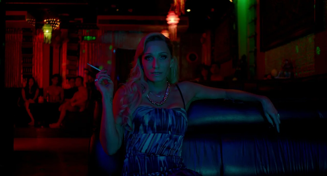 Only God Forgives (2013) movie still: medium — A dark lounge area features deep shadows contrasted by intense red and green light. A blonde woman…