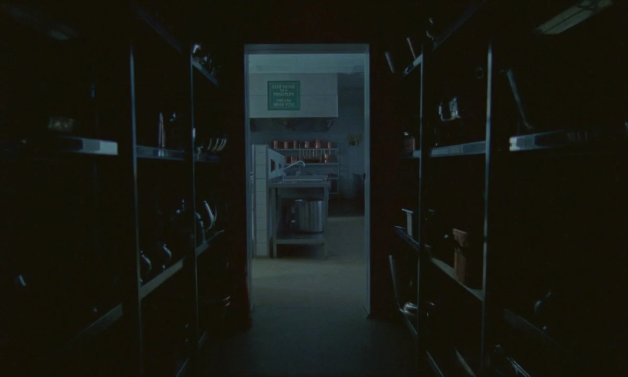 Spencer (2021) movie still: establishing — A dark, narrow metal shelving unit frames the entrance to a sterile prep area. A green instructional sign…