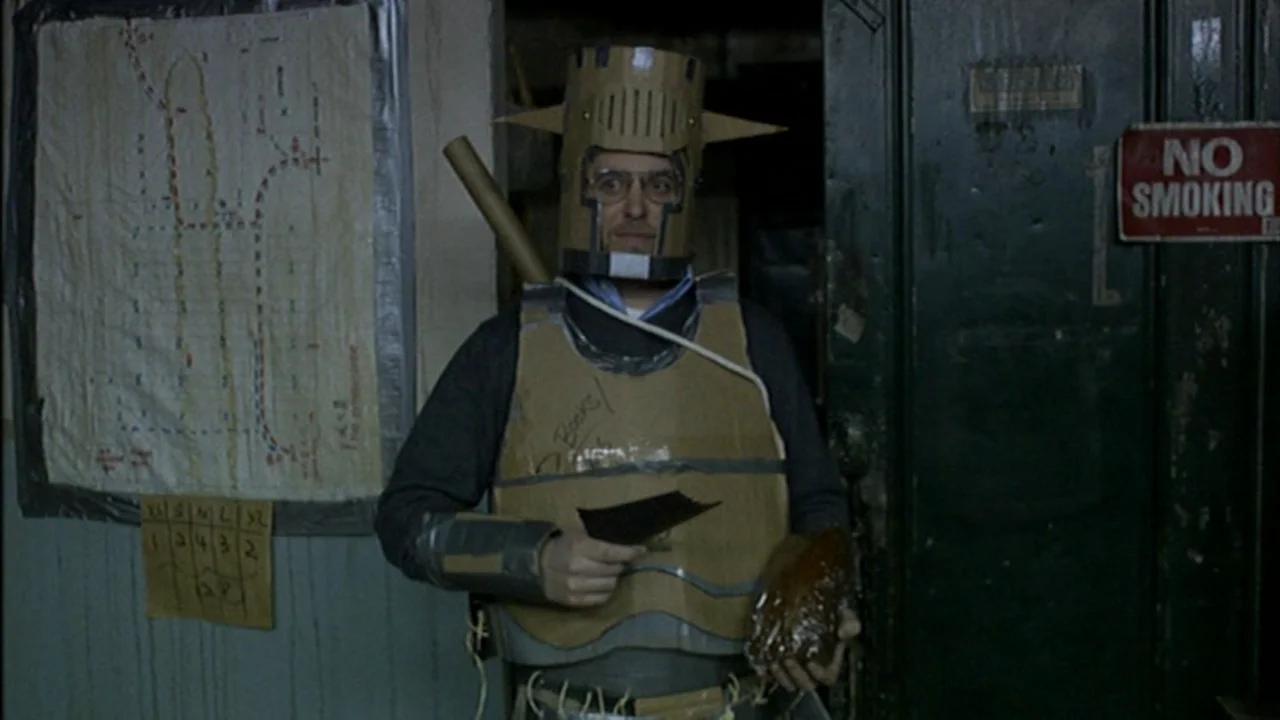 Murder Party movie still: medium — A dimly lit utility room features a wall map and a no smoking sign. A man wears homemade cardboard armor and…