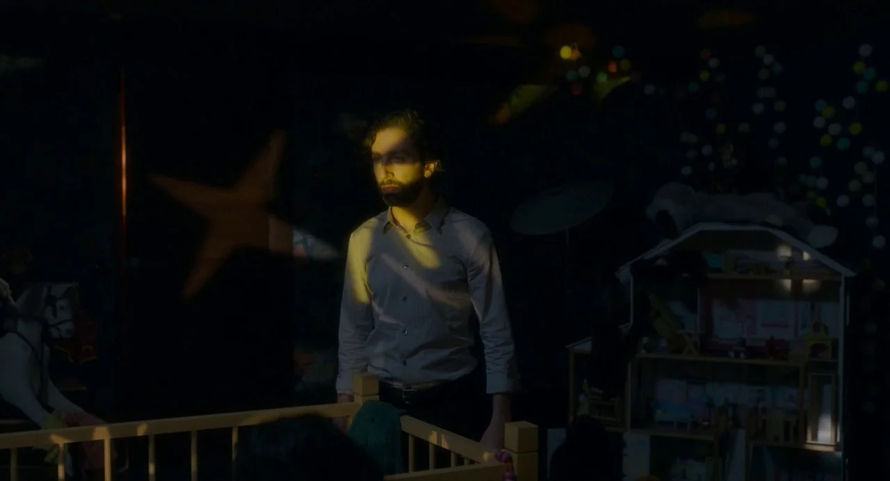 Annette (2021) movie still: medium — A dimly lit room filled with toys and a wooden baby crib. A man stands in the darkness while a projected star…