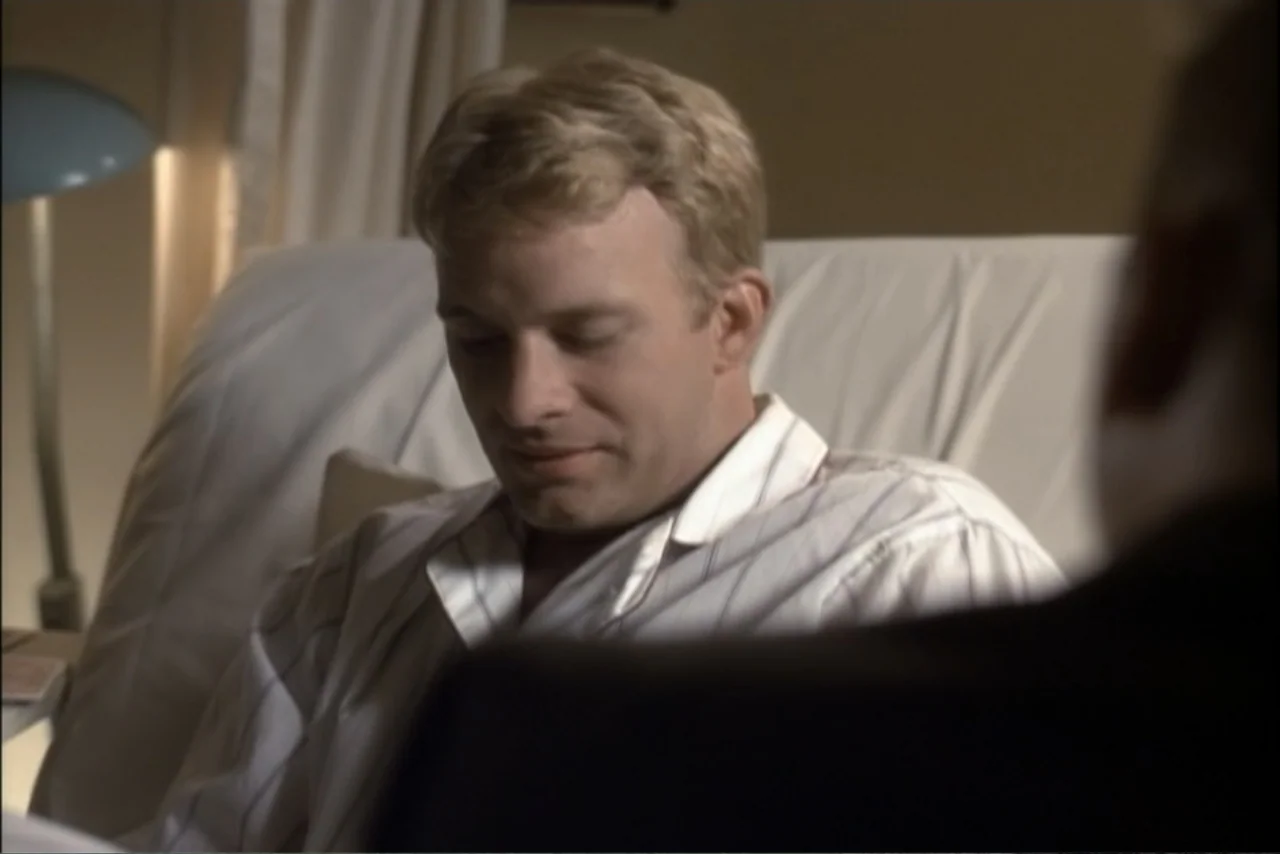 Unification of Japan 61 (2024) movie still: medium — A man sits in a hospital bed with white linens and a nearby lamp. He wears pajamas and looks…