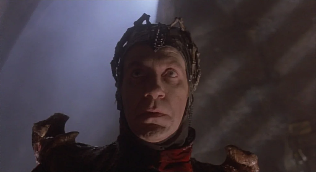 Time Bandits (1981) movie still: closeup — A dark stone-like interior with minimal detail visible behind a person. The man wears a skeletal…
