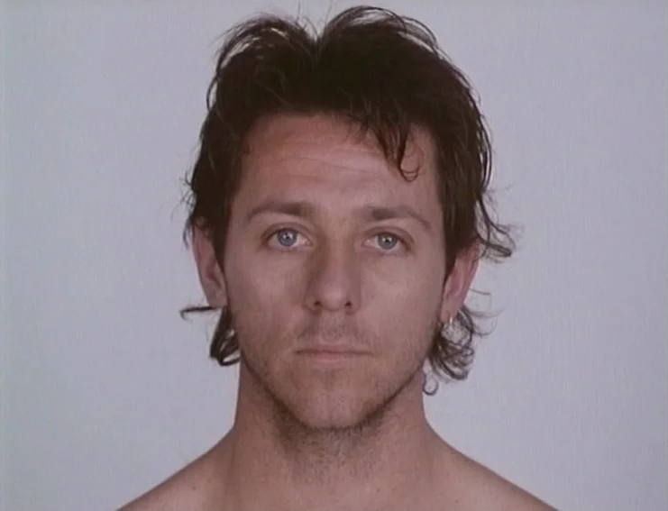 Ghosts… Of The Civil Dead (1988) movie still: closeup — A man stands centered against a flat, neutral light gray backdrop. The shot is a direct,…
