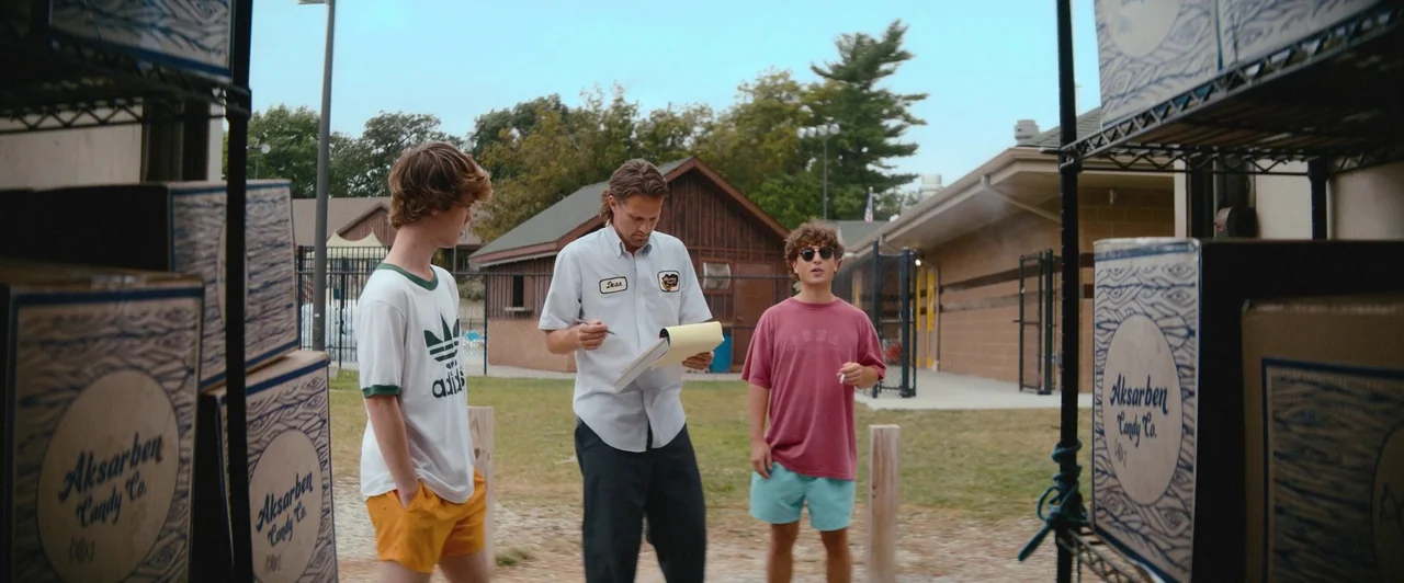 Snack Shack (2024) movie still: fullbody — An outdoor area features stacked cardboard shipping boxes near a brick service building. Three men stand…