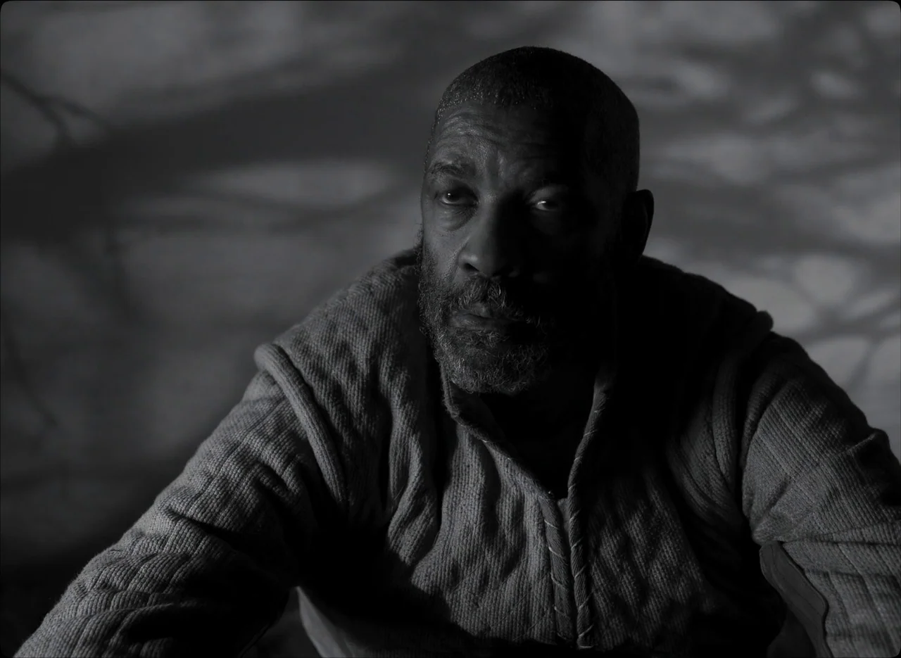 The Tragedy of Macbeth (2021) movie still: medium — A man sits in a dimly lit space with patterned shadows cast behind him. The person wears a…