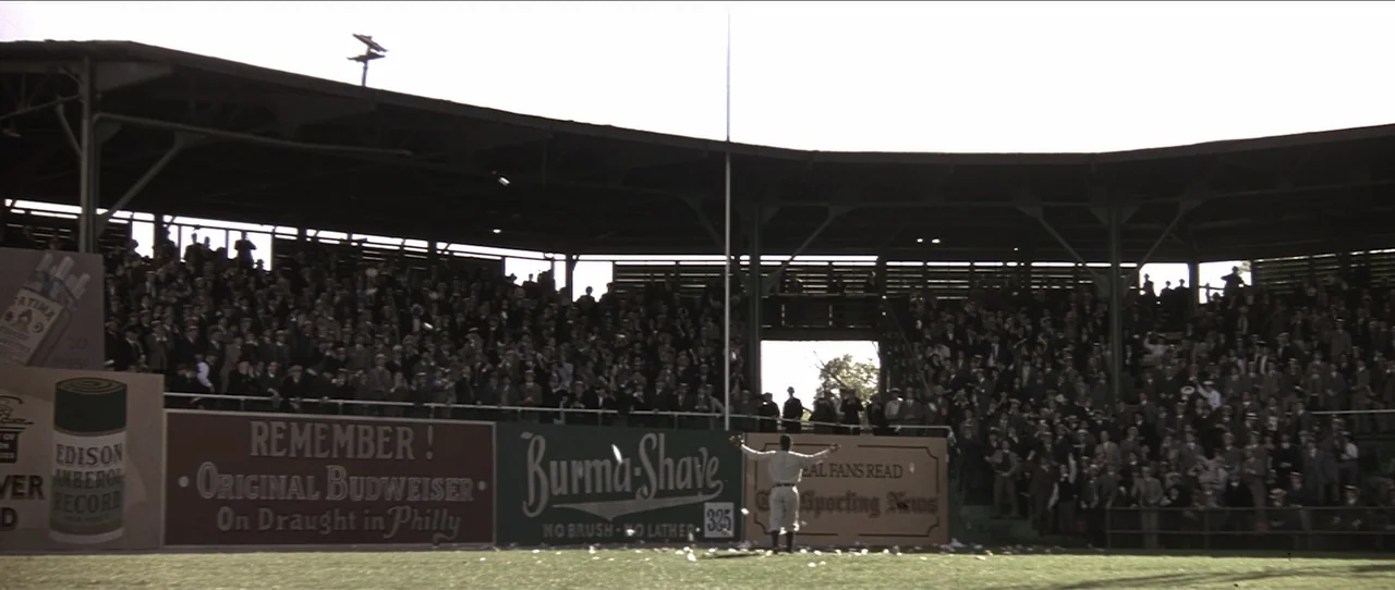 Cobb (1994) movie still: wide — A historic wooden baseball stadium is filled with a large crowd in period attire. A lone baseball player in a white…
