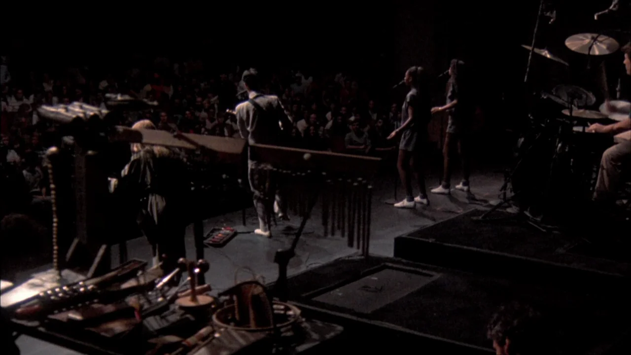 Stop Making Sense (1984) movie still: wide — A dimly lit concert hall stage features a band performing for an unseen audience. The musician in a…