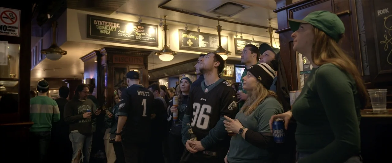 Kelce (2023) movie still: medium — A crowded sports bar interior features patrons wearing football jerseys under low hanging light fixtures. Several…