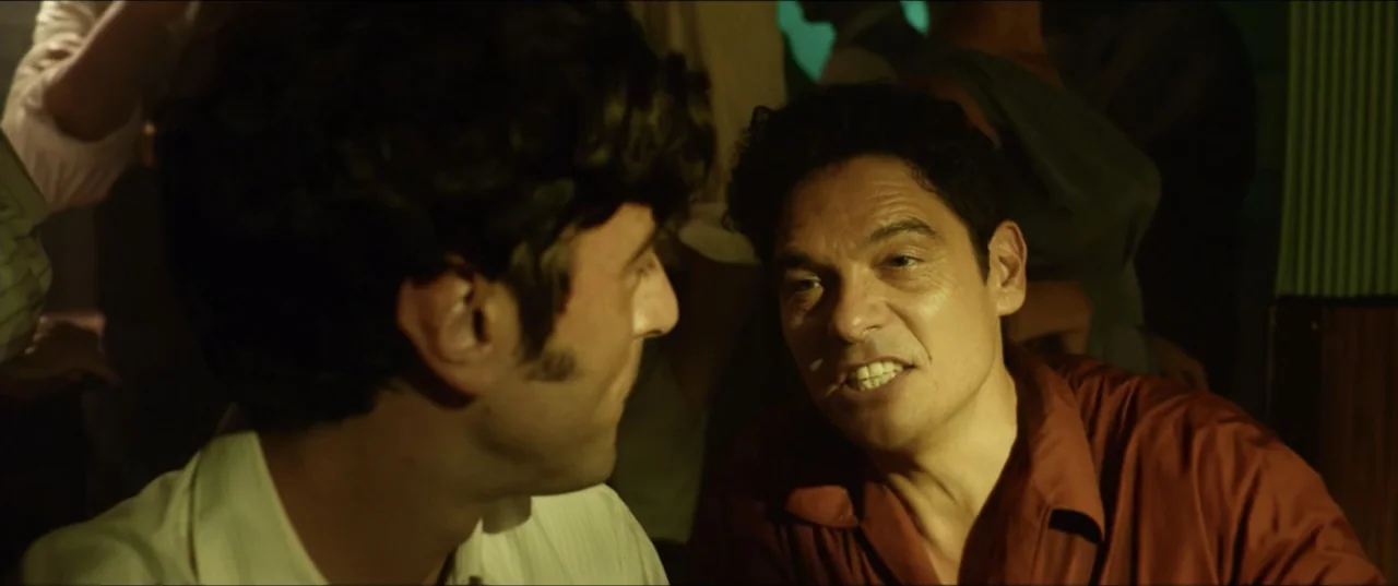 Hear Me: Our Summer (2024) movie still: medium — A dimly lit, crowded indoor bar setting with visible silhouettes in the background. A man in a…