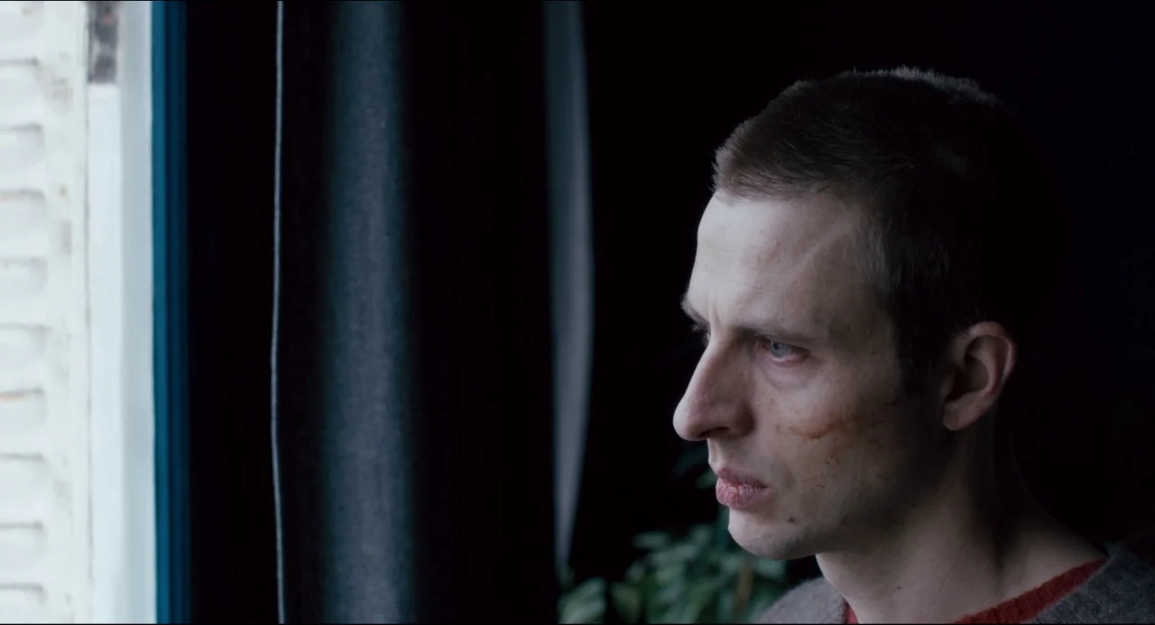 The Night Eats the World (2018) movie still: closeup — A close-up of a man standing in a dimly lit room near a window. He stares out the window with…