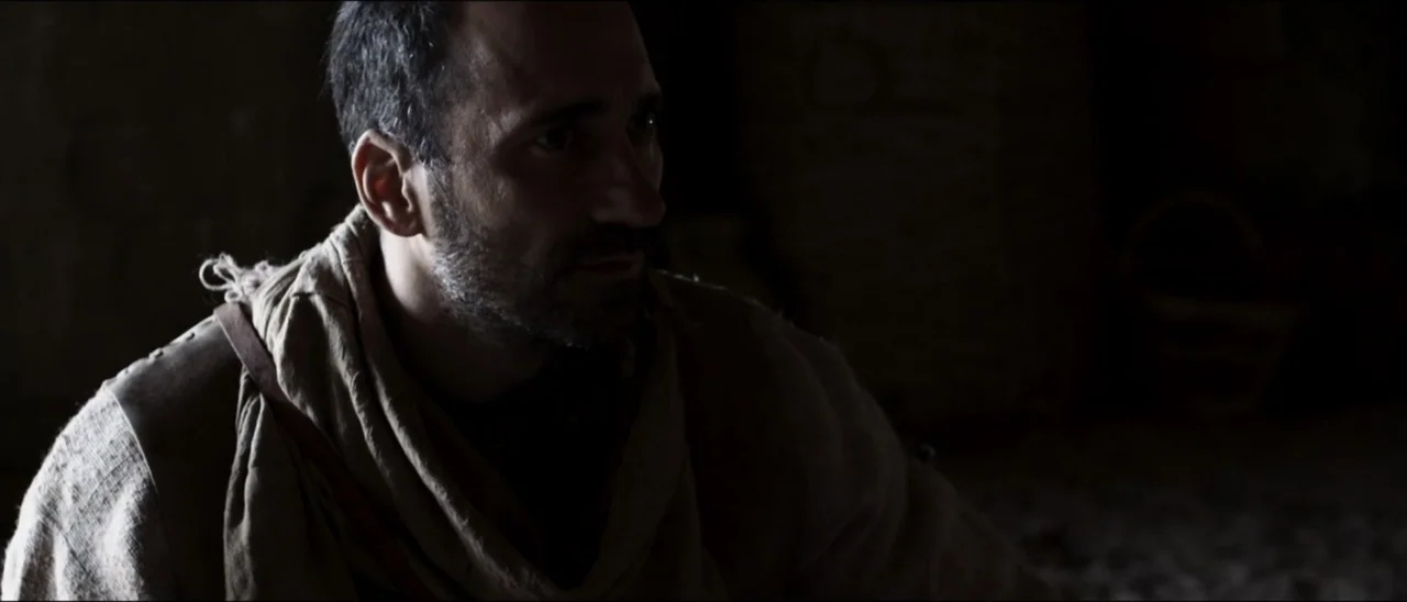 The Penitent Thief (2021) movie still: medium — A dimly lit interior space with shadows obscuring the surroundings. A man with short dark hair gazes…