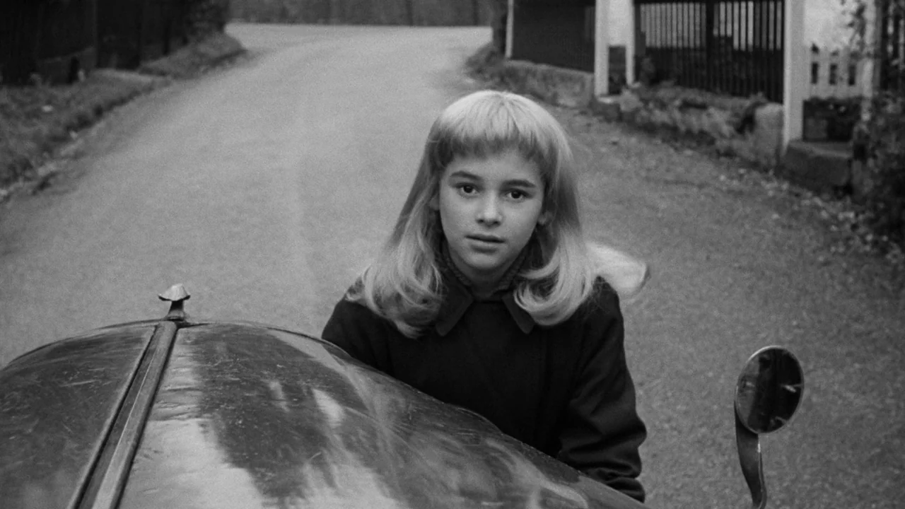 Village of the Damned (1960) movie still: medium — A quiet paved road winds through a rural area with a fence visible nearby. A young blonde girl…