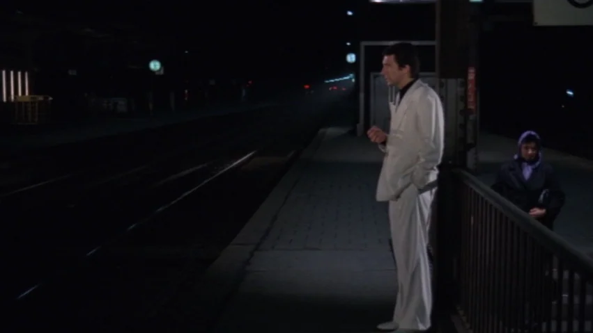 Tenderness of the Wolves (1973) movie still: fullbody — A dark, deserted outdoor train platform at night with distant flickering lights. A man in a…