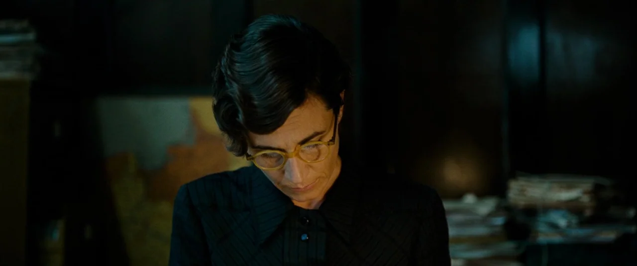 Phoenix (2014) movie still: closeup — A dark, cluttered office setting features stacks of paper visible in the blurred background. A woman with dark…