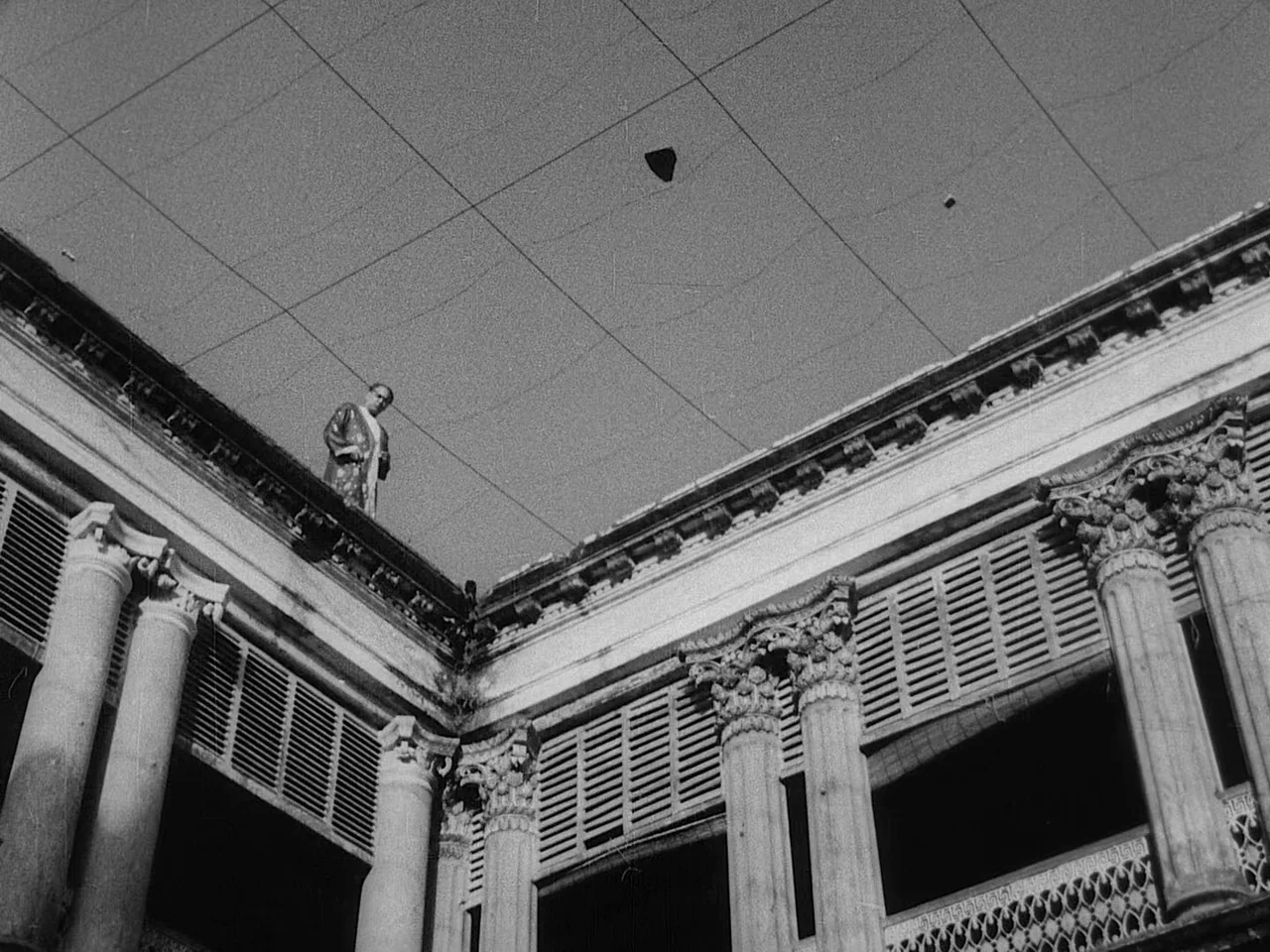 The Music Room (1958) movie still: wide — A dilapidated courtyard features neoclassical columns and decorative balustrades under a cloudy sky. A man…