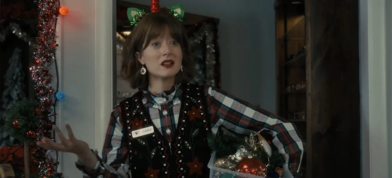 Merv (2025) movie still: medium — A festive indoor environment decorated with tinsel and Christmas baubles on display shelves. A woman wearing a…
