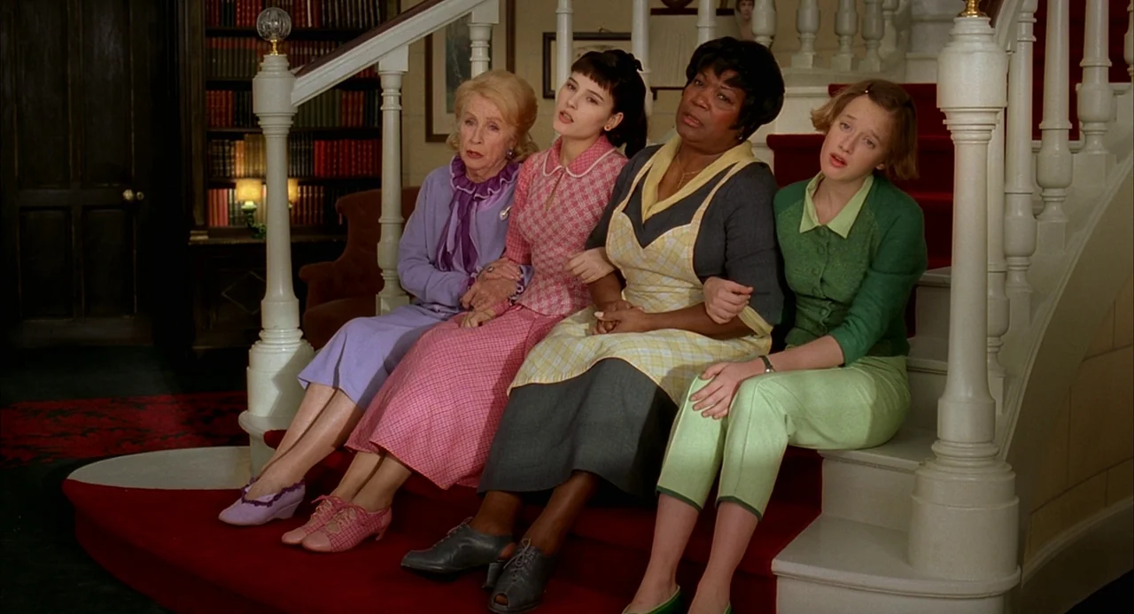 8 Women (2002) movie still: fullbody — A group of four women sits on a red-carpeted staircase in a large home. They sit in a horizontal row looking…