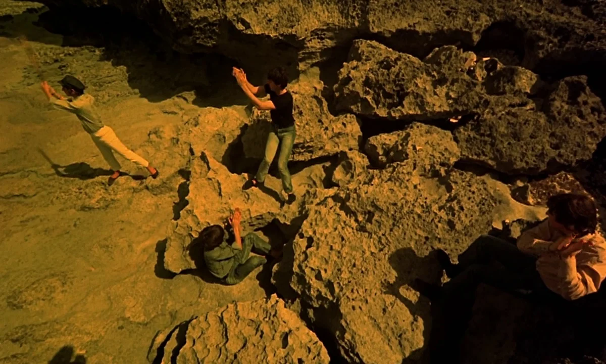 Help! (1965) movie still: wide — A rugged beach area features porous, uneven rock formations scattered across the ground. Four men are positioned…