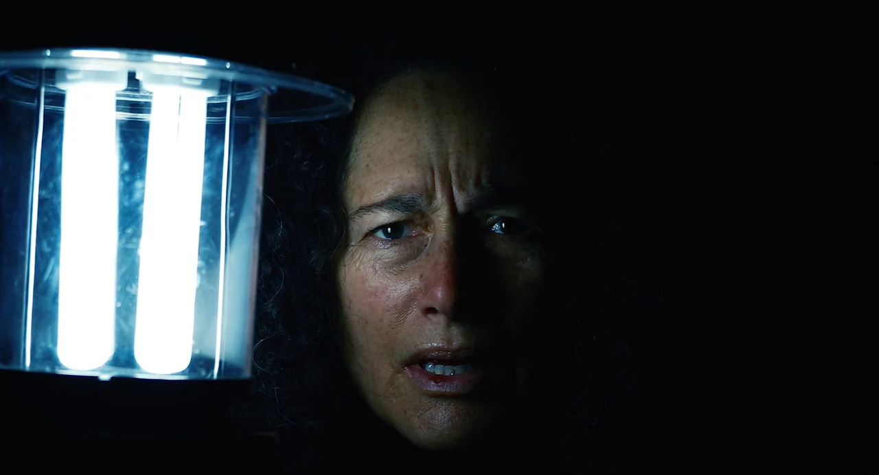 Hellbender (2021) movie still: closeup — A pitch-black room is illuminated only by a portable fluorescent camping lantern. A middle-aged woman is…