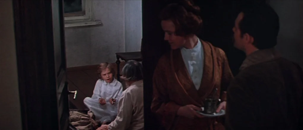Dersu Uzala (1975) movie still: medium — A dimly lit room with wooden floors is viewed from a dark doorway. A woman holds a small cup and saucer…