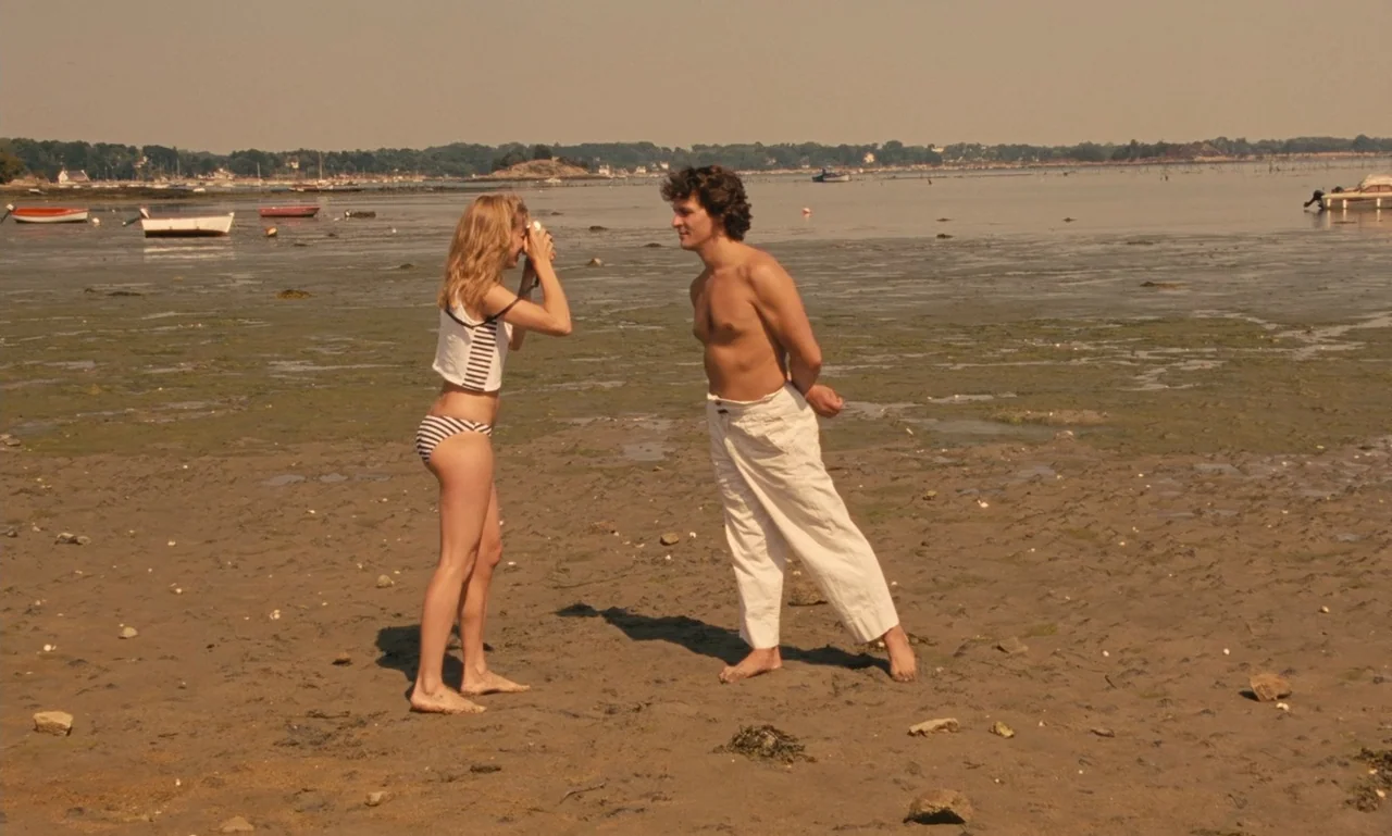 A Tale of Winter (1992) movie still: fullbody — A low-tide beach with exposed mudflats and several small docked boats in the distance. A woman…