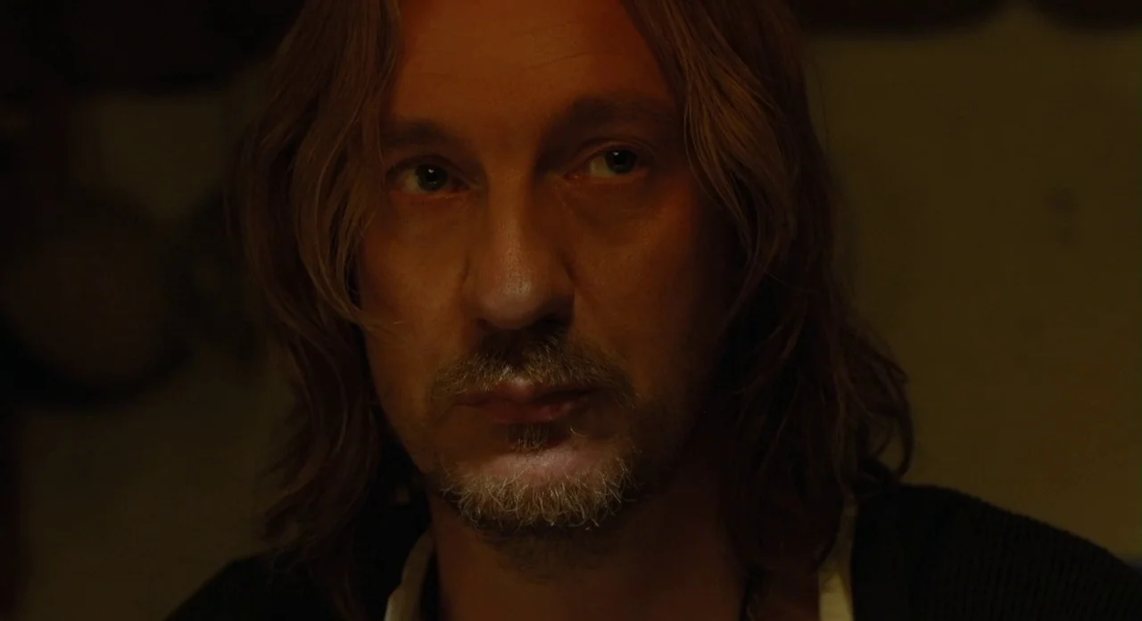 London Boulevard (2010) movie still: closeup — A dimly lit room with muted earth tones and deep shadows in the background. A man with long brown…