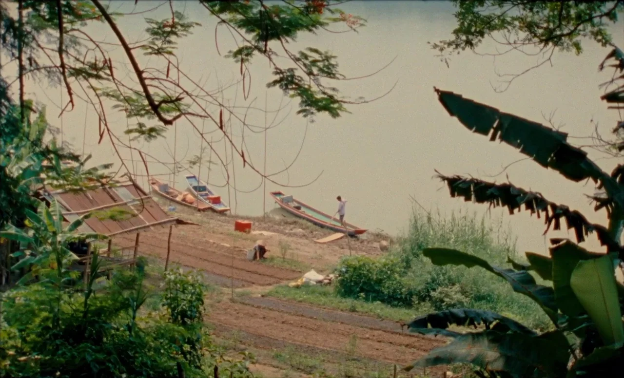 Samsara (2023) movie still: wide — A rural riverbank features tilled garden rows, wooden boats, and sparse tropical vegetation. One person stands in…