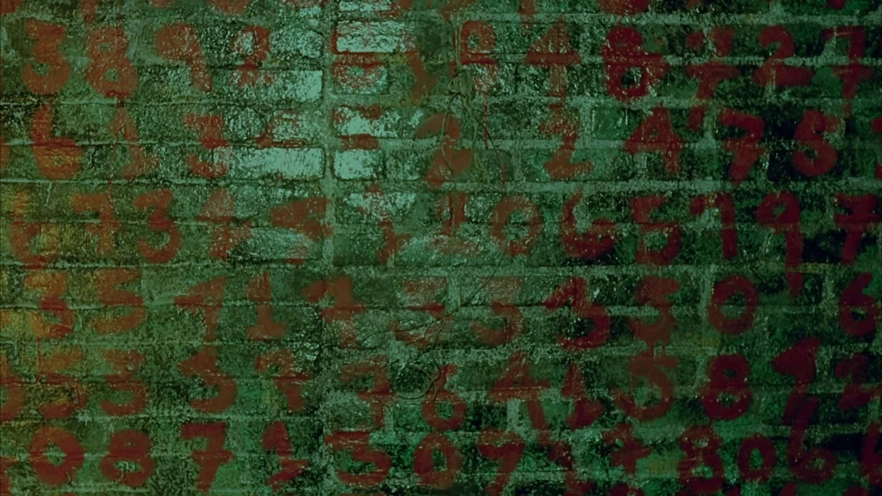 Saw movie still: closeup — A dark, moisture-slicked brick wall is covered in repeated, hand-painted red digits. The surface shows signs of age and…