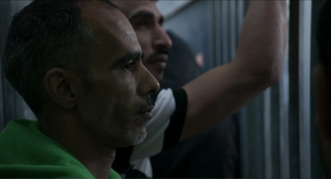 Human Flow (2017) movie still: medium — A dark, cramped space features vertical grey metal bars in the foreground. Two men stand behind the bars…
