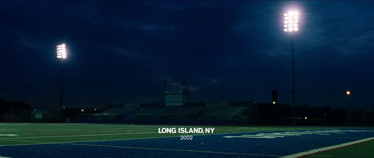 Bad Education (2019) movie still: establishing — A quiet high school athletic stadium at night under bright overhead floodlights. The artificial…