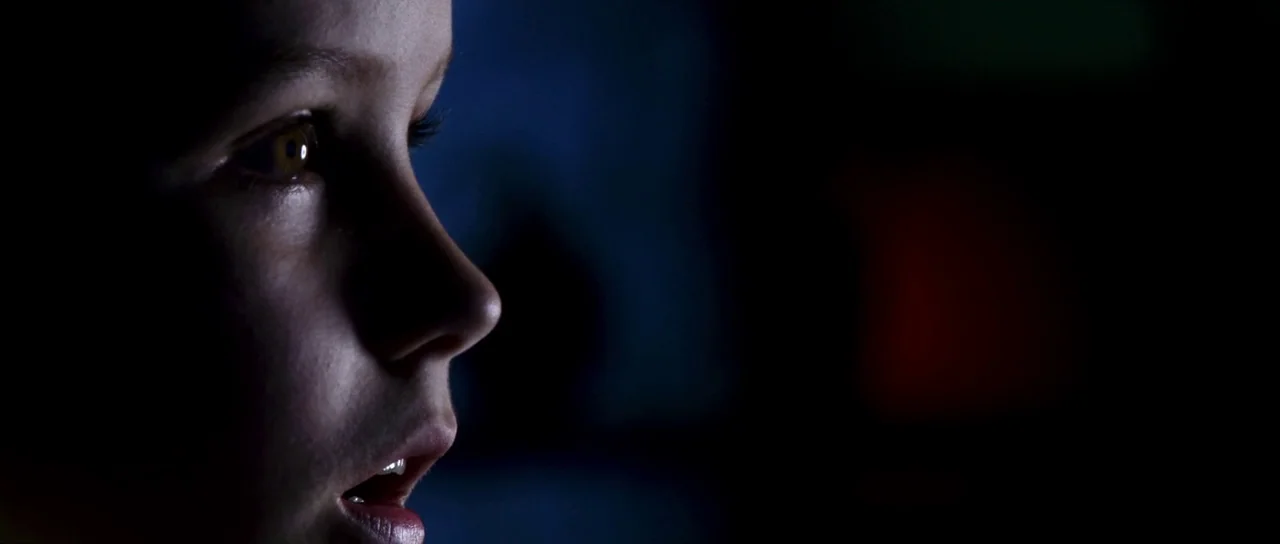 Speed Racer (2008) movie still: closeup — A dark, unlit room provides a stark black backdrop to a single child face. The child stares intently at a…