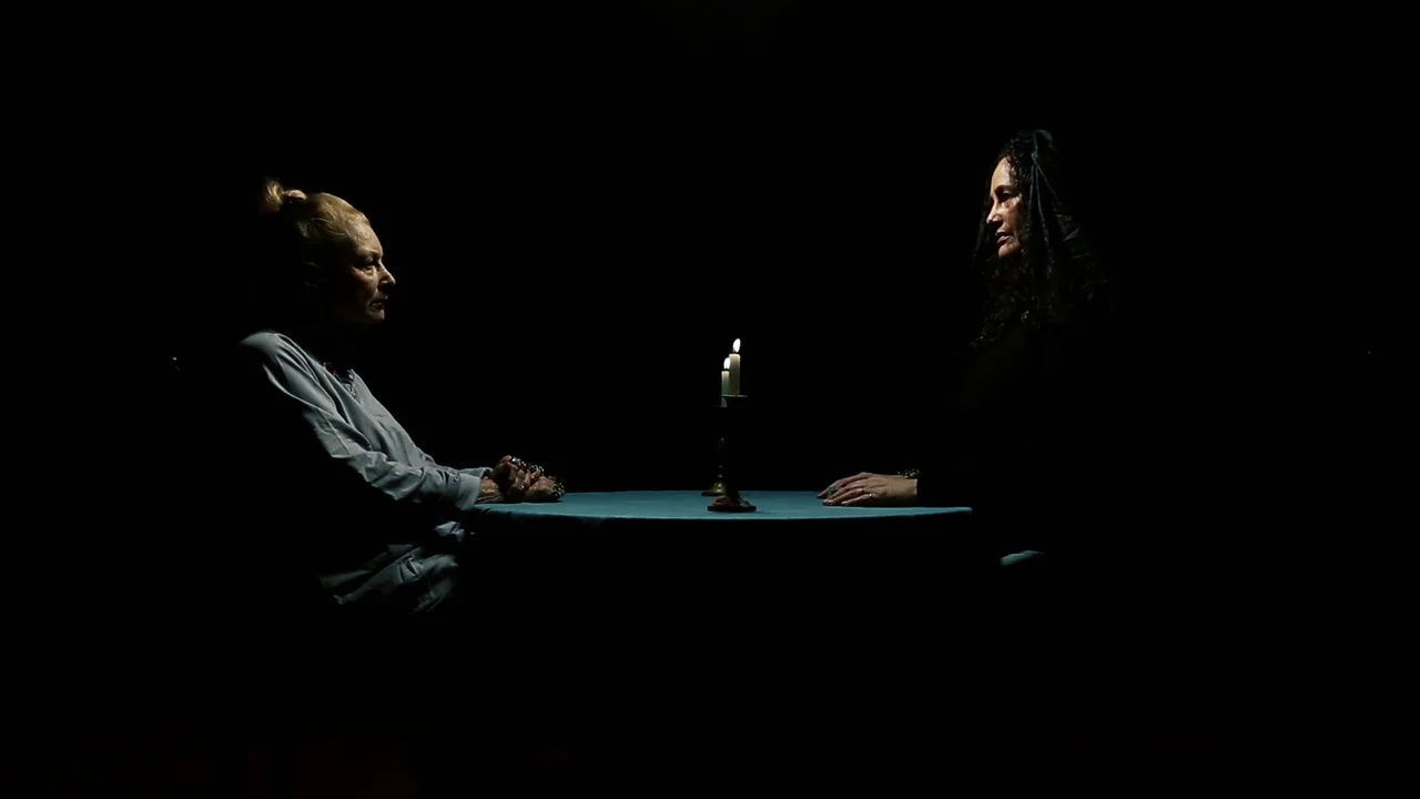 The Deeper You Dig (2019) movie still: medium — A dark space features two women sitting at a small, round table facing each other. A single lit…
