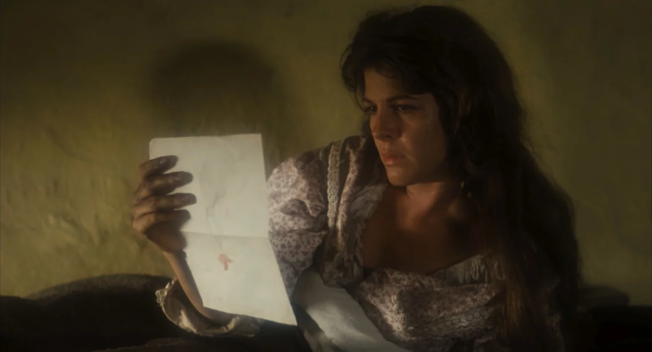 Camila (1984) movie still: medium — A dark, minimally lit interior room with a textured wall. A young woman reads a handwritten letter while sitting…