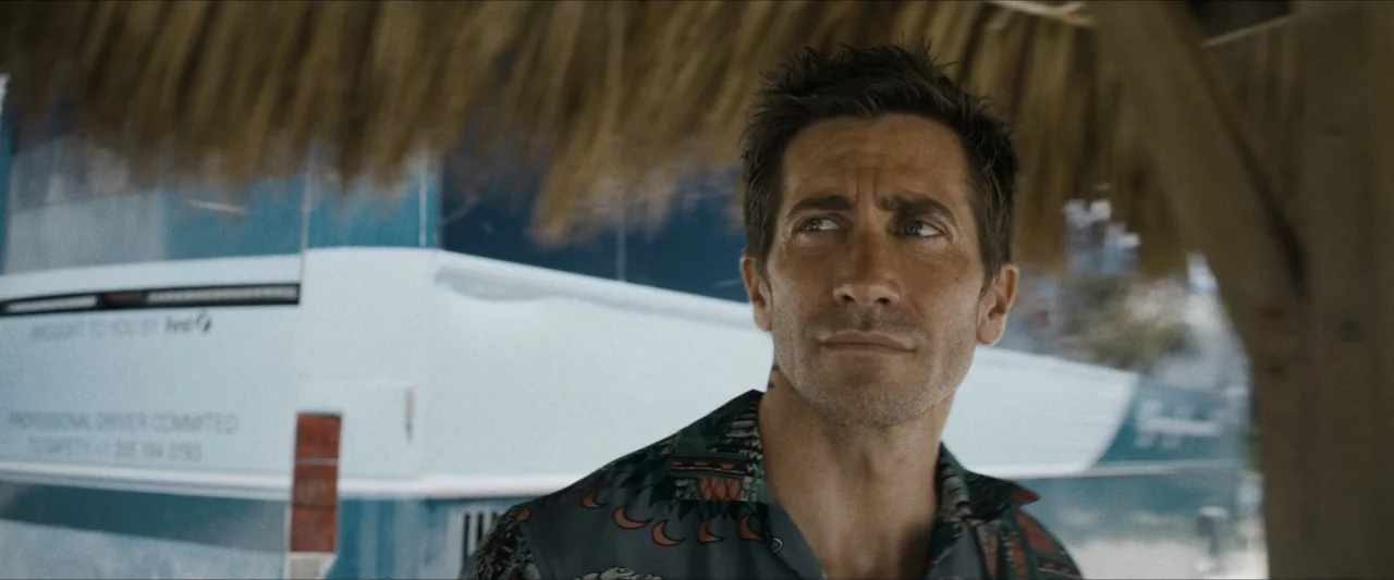 Road House (2024) movie still: medium — An outdoor area with a thatched roof covering a space near a parked vehicle. A man stands under the shade,…