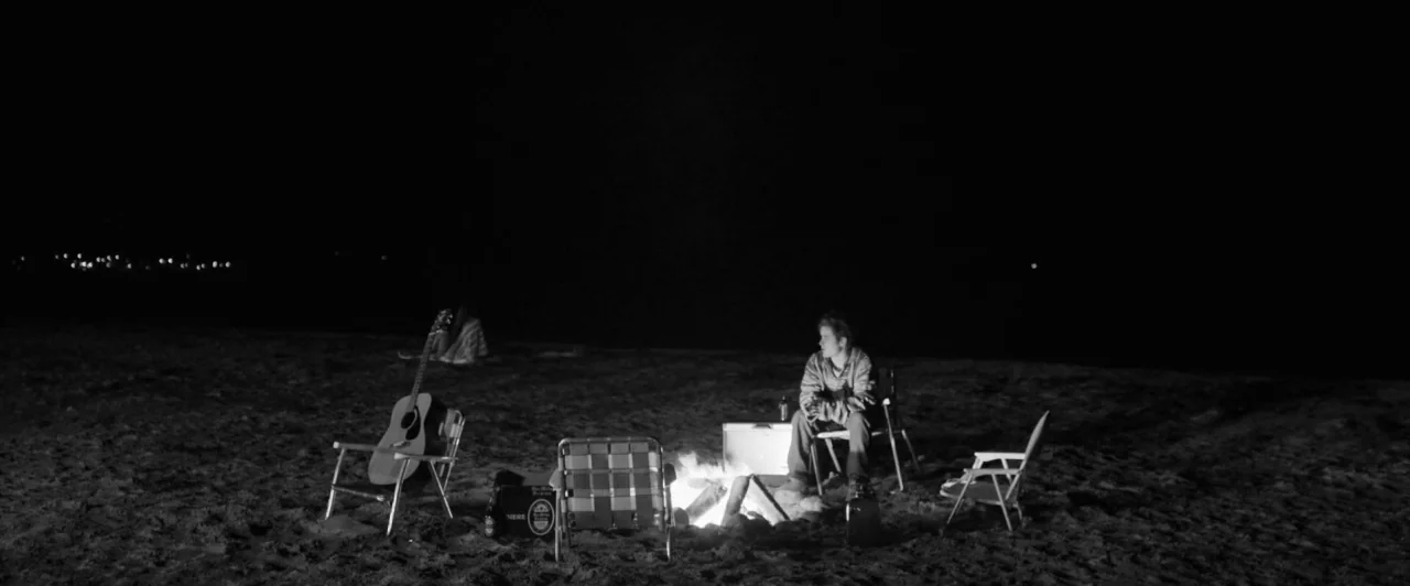 Weirdos (2016) movie still: wide — A dark, empty beach at night is lit by a small, central campfire. A young man sits in a folding chair while an…