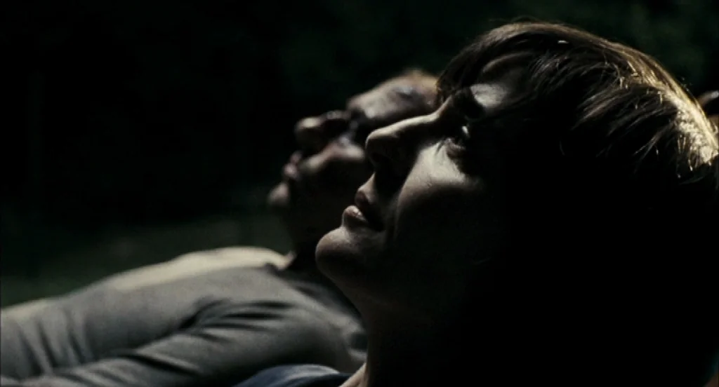 Timecrimes (2007) movie still: closeup — Two individuals lie side-by-side on the ground in a dark outdoor setting at night. They both gaze upwards…