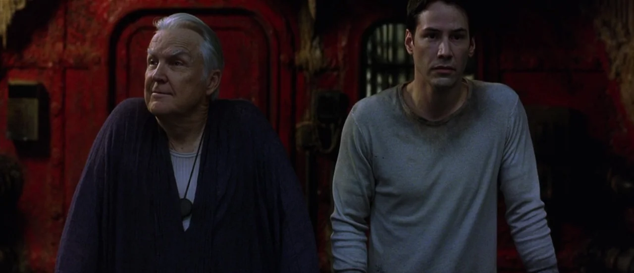 The Matrix Reloaded (2003) movie still: medium — An interior space with distressed red metal plating on the rear wall. An elderly man and a younger…