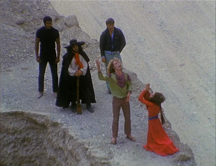 The Niklashausen Journey (1970) movie still: wide — A group of people stands on the edge of a steep rocky desert cliff. One blonde man gestures…
