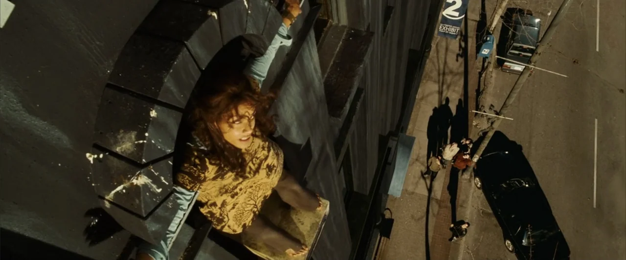 Catwoman (2004) movie still: medium — A woman stands on a narrow ledge outside a tall building in an urban setting. She looks directly up at the…