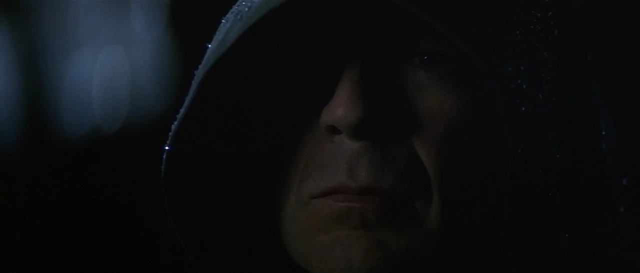 Unbreakable (2000) movie still: closeup — A dark outdoor setting at night features a man wearing a hooded jacket. The subject stands in shadow with…