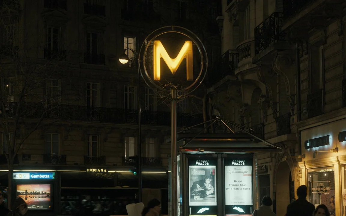 Let The Sunshine In (2017) movie still: establishing — A Parisian street at night features a prominent yellow backlit subway entrance sign. Stone…