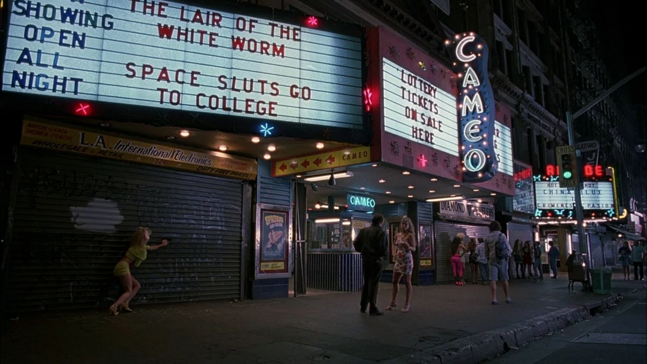 Whore (1991) movie still: wide — A night city sidewalk features storefronts with illuminated marquees for a vintage theater. A small group of people…