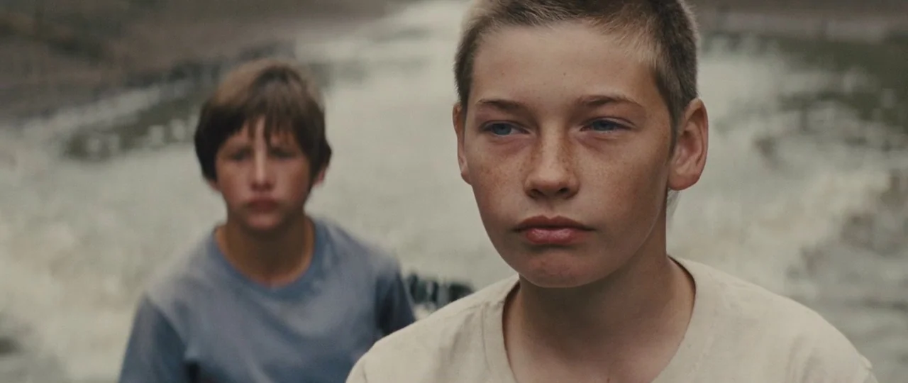 Mud (2012) movie still: medium — Two teenage boys stand in the foreground against a backdrop of moving water. The focus is sharp on the boy in front…