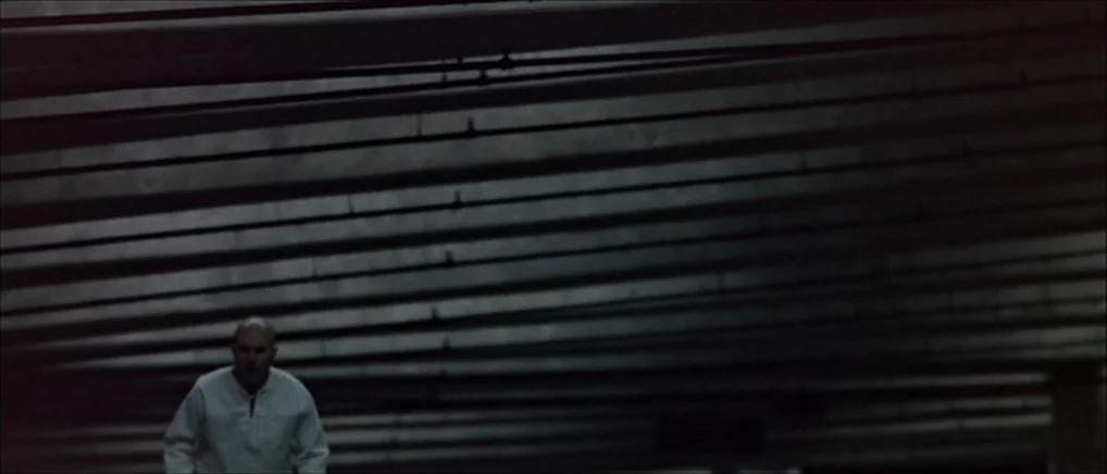 THX 1138 (1971) movie still: wide — A dark, expansive parking garage features prominent parallel concrete support beams overhead. A lone bald man in…