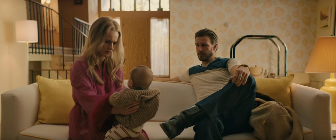 I'm Your Woman (2020) movie still: medium — A vintage 1970s living room features floral patterned wallpaper and a cream-colored sofa. A blonde woman…